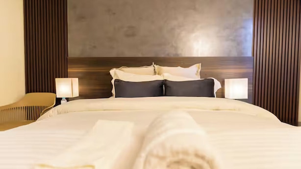 A hotel room with a large bed, two bedside lamps, a wooden headboard, and a rolled white towel on the bed.