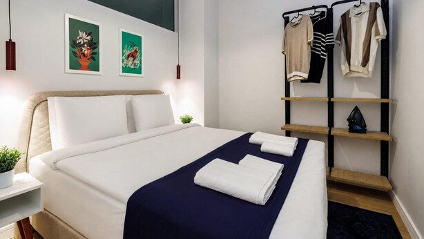 A modern hotel room with a neatly made bed, a bedside table with a plant, a wall-mounted shelf with clothes, and two framed artworks. - Fantastic Cihangir 3 Bedroom Apt At Monnas Suite (Istanbul)