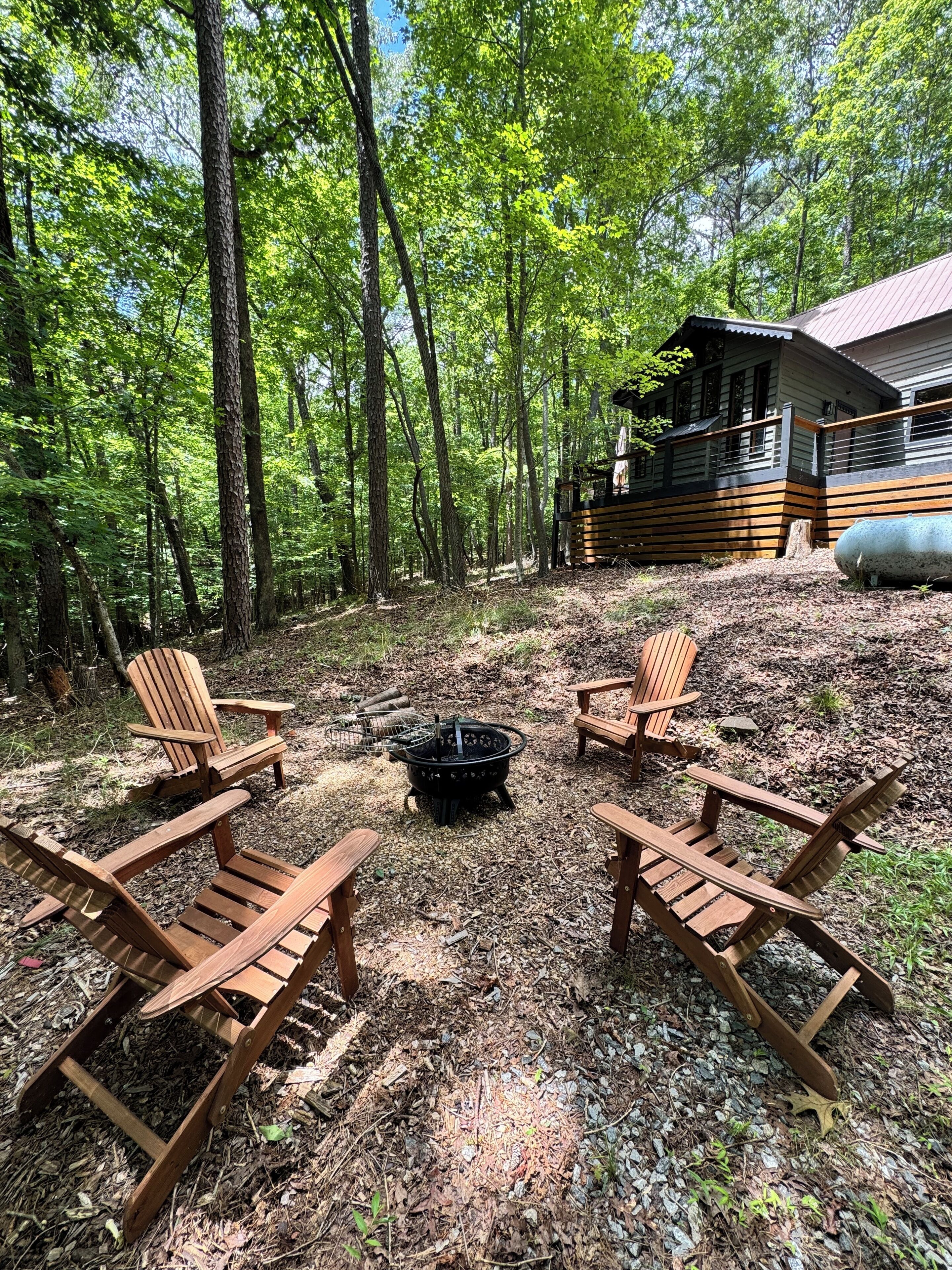 A cabin with a deck, surrounded by a forest. There are four Adirondack chairs and a fire pit in the clearing.