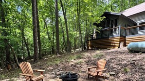 A cabin with a deck, surrounded by a forest. There are four Adirondack chairs and a fire pit in the clearing.