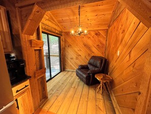 Interior acolhedor de uma cabana com teto de madeira, uma poltrona de couro, um banquinho de madeira e uma cafeteira.
