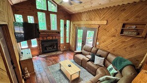 A wooden cabin interior with a fireplace, a sofa, a television, and large windows.