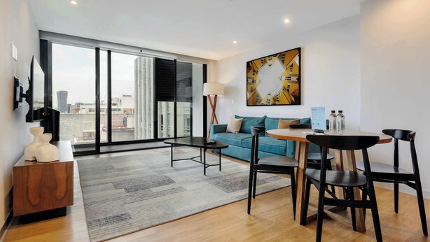 A modern living room with a sofa, a dining table with chairs, a large window, and a painting on the wall. - w* | Upscale 1BR w/ AC in the Heart of Roma Sur (Ciudad de México)