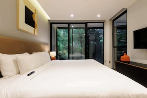 Ein modernes Hotelzimmer mit einem großen Bett, einem Flachbildfernseher und einer Glastür, die auf einen Balkon mit Blick auf Bäume führt.