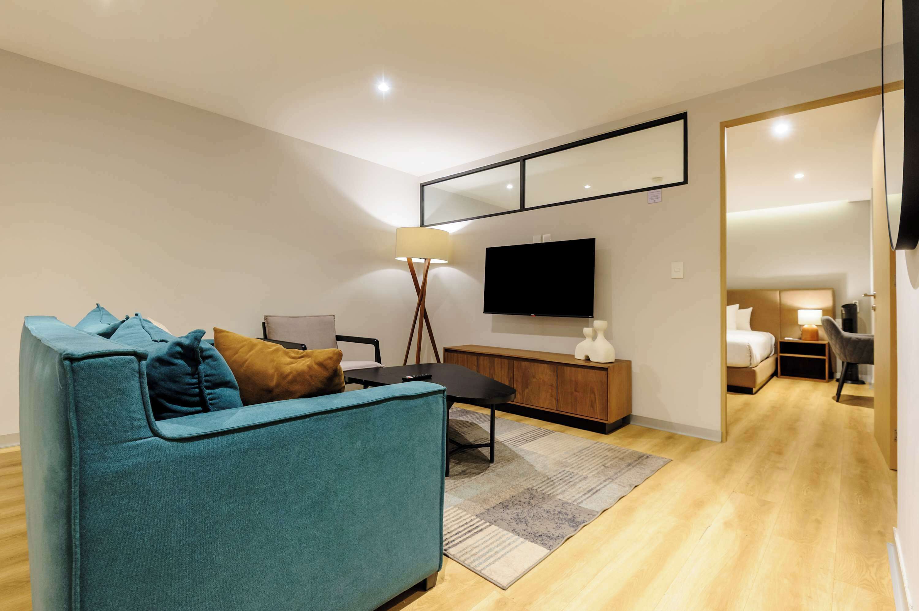A modern living room with a teal sofa, a flat-screen TV, a wooden cabinet, and a mirror on the wall.
