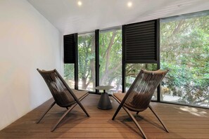 Ein Zimmer mit zwei Korbstühlen, einem kleinen runden Tisch und großen Fenstern mit schwarzen Rollläden.