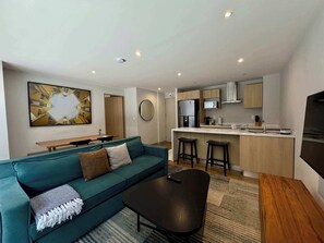 Ein modernes Wohnzimmer mit einem petrolfarbenen Sofa, einem Couchtisch und einer Küchenzeile mit Kühlschrank, Mikrowelle und Barhocker.