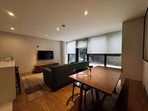 Ein modernes Wohnzimmer mit einem Holzesstisch, einem dunkelgrünen Sofa, einem Flachbildfernseher und einer Küchenzeile mit Spüle.