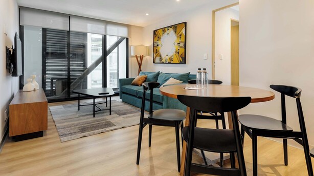 A modern living room with a sofa, a dining table with chairs, a coffee table, a lamp, and a large window with blinds. - w* | Stunning 1BR w/ AC in the Heart of Roma Sur (Ciudad de México)