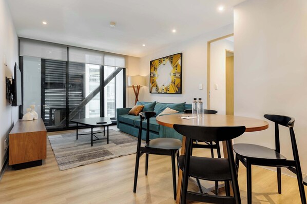 A modern living room with a sofa, a dining table with chairs, a coffee table, a lamp, and a large window with blinds. - w* | Stunning 1BR w/ AC in the Heart of Roma Sur (Ciudad de México)