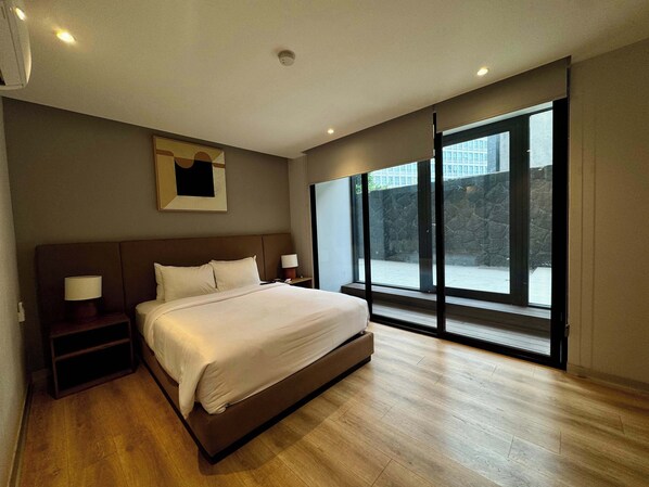 Ein modernes Schlafzimmer mit einem großen Bett, Nachttischlampen, einem Beistelltisch, einem Fenster mit Blick auf eine Steinmauer und ein Gebäude sowie eingelassener Deckenbeleuchtung.