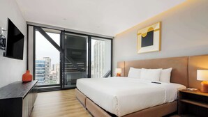 Ein modernes Hotelzimmer mit einem großen Bett, einem Schreibtisch, einem Fernseher und Blick auf die Stadtlandschaft durch eine Glastür.