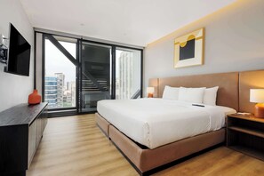 Ein modernes Hotelzimmer mit einem großen Bett, einem Schreibtisch, einem Fernseher und Blick auf die Stadtlandschaft durch eine Glastür.