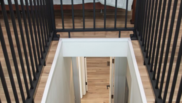 Una escalera moderna con barandillas negras y peldaños de madera que conduce a un dormitorio con cama blanca y suelo de madera.
