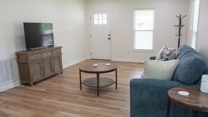 Una sala de estar con una televisión de pantalla plana, un mueble de madera, un sofá azul, una mesa redonda y un perchero.