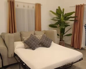 Ein Bett mit weißer Matratze und zwei gemusterten Kissen, ein beiges Sofa, eine Topfpflanze und Vorhänge in einem Zimmer.