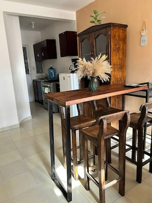 A dining area with a wooden table and chairs, a vase with dried plants, and a kitchen in the background.