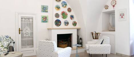 A room with two white armchairs, a small table, a fireplace, and a wall decorated with colorful plates.