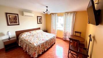 A bedroom with a bed, a bedside table, a chair, a TV, and a window with curtains.