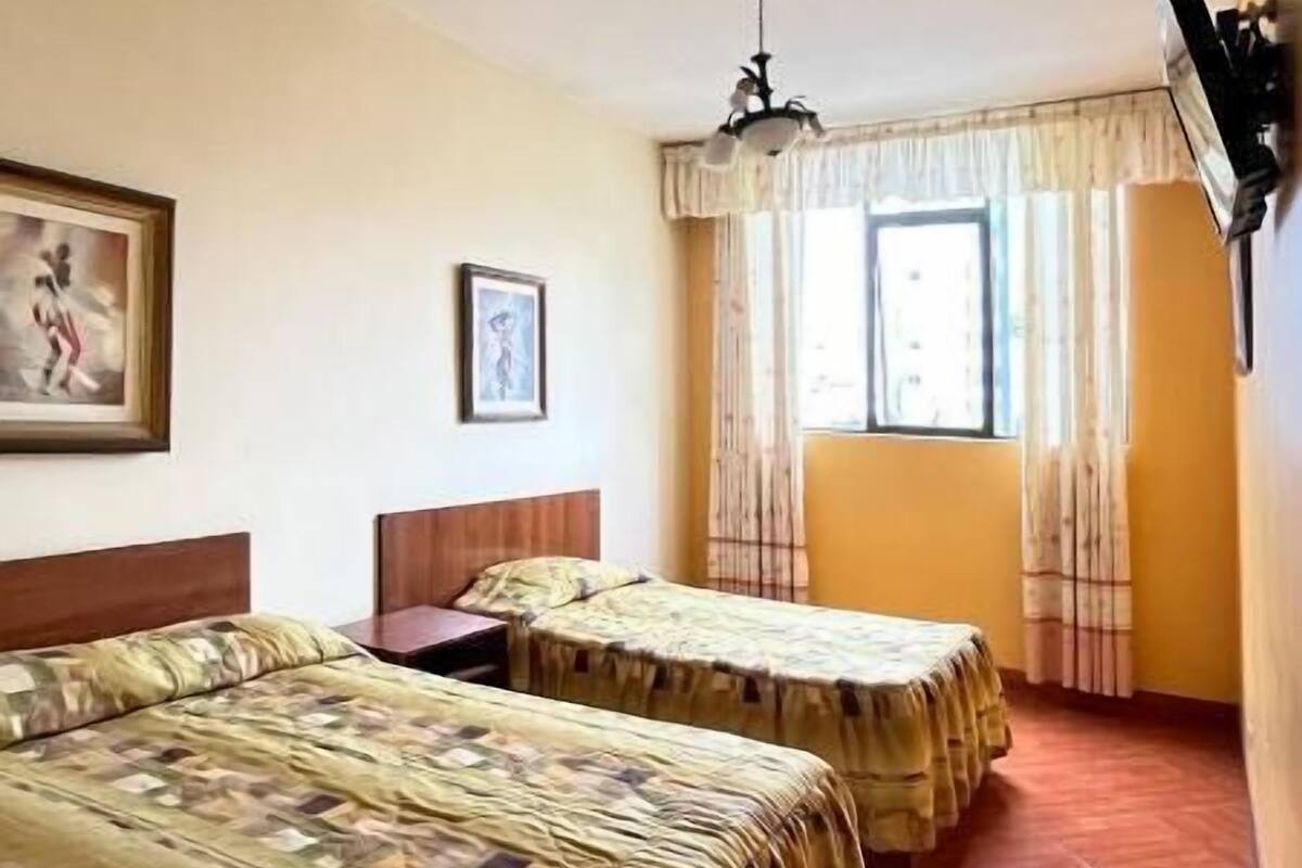 Habitación de hotel con dos camas, ventilador de techo, ventana con cortinas y televisor montado.