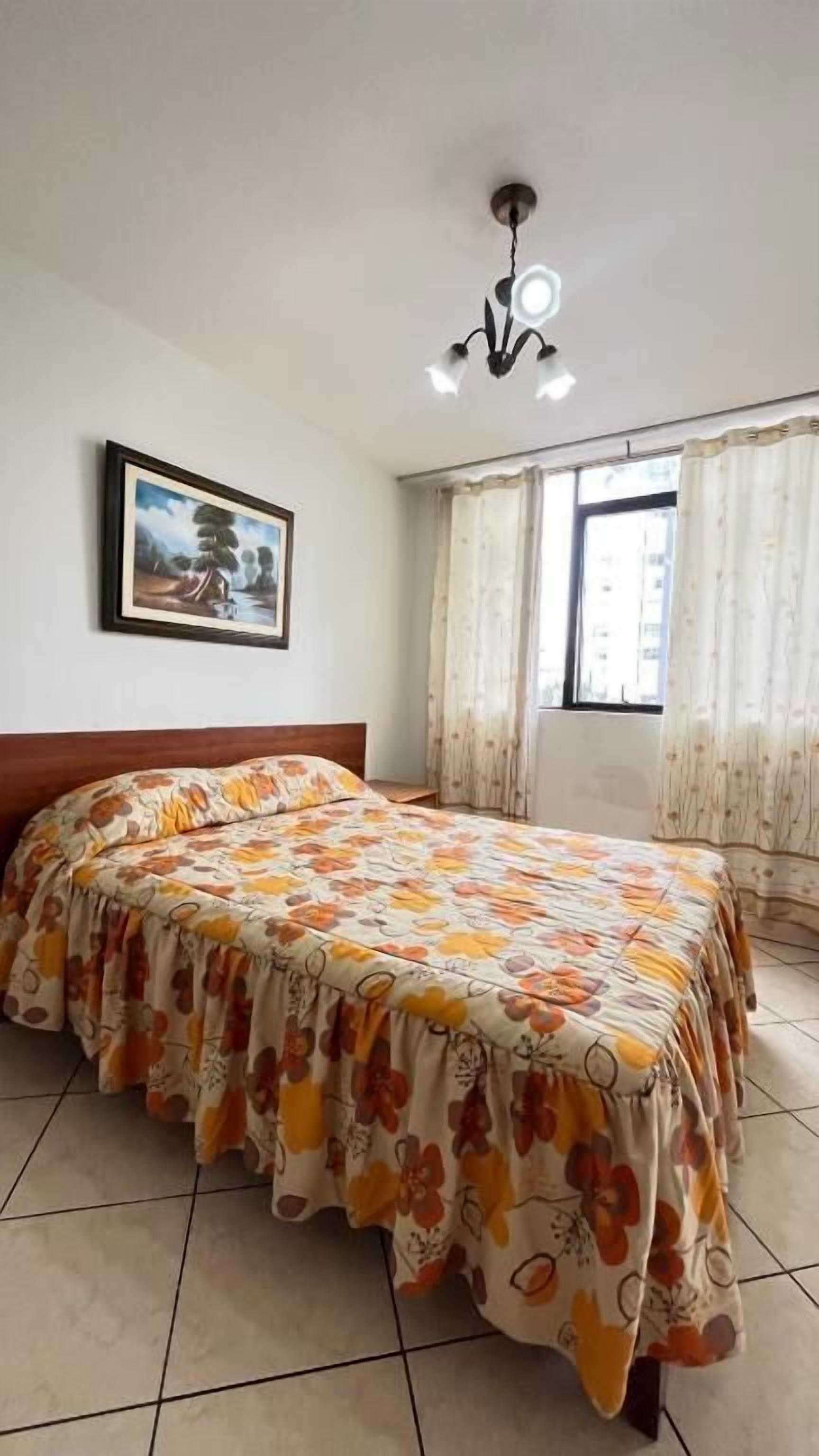 A bedroom with a bed, curtains, a painting, and a ceiling light fixture.