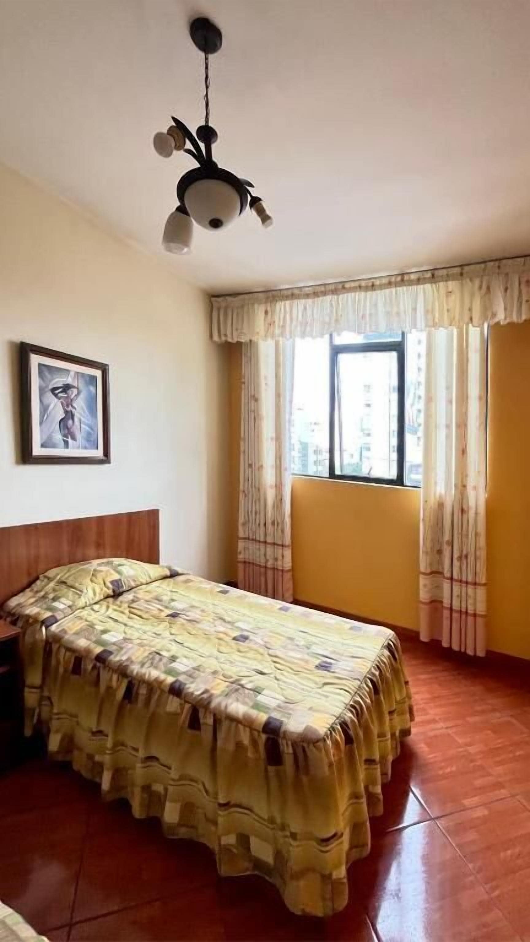 Quarto com cama, janela com cortinas, quadro na parede e luminária no teto.