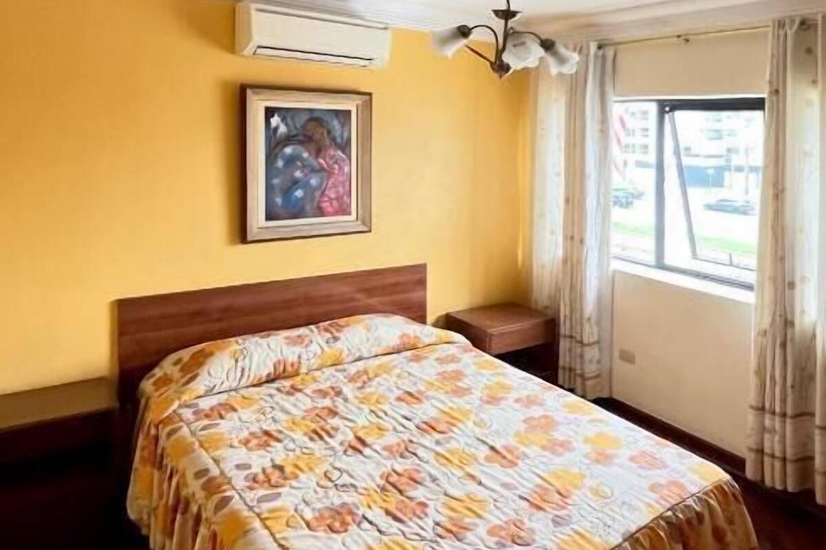 Un dormitorio con cama, ventilador de techo, ventana con cortinas y aire acondicionado.