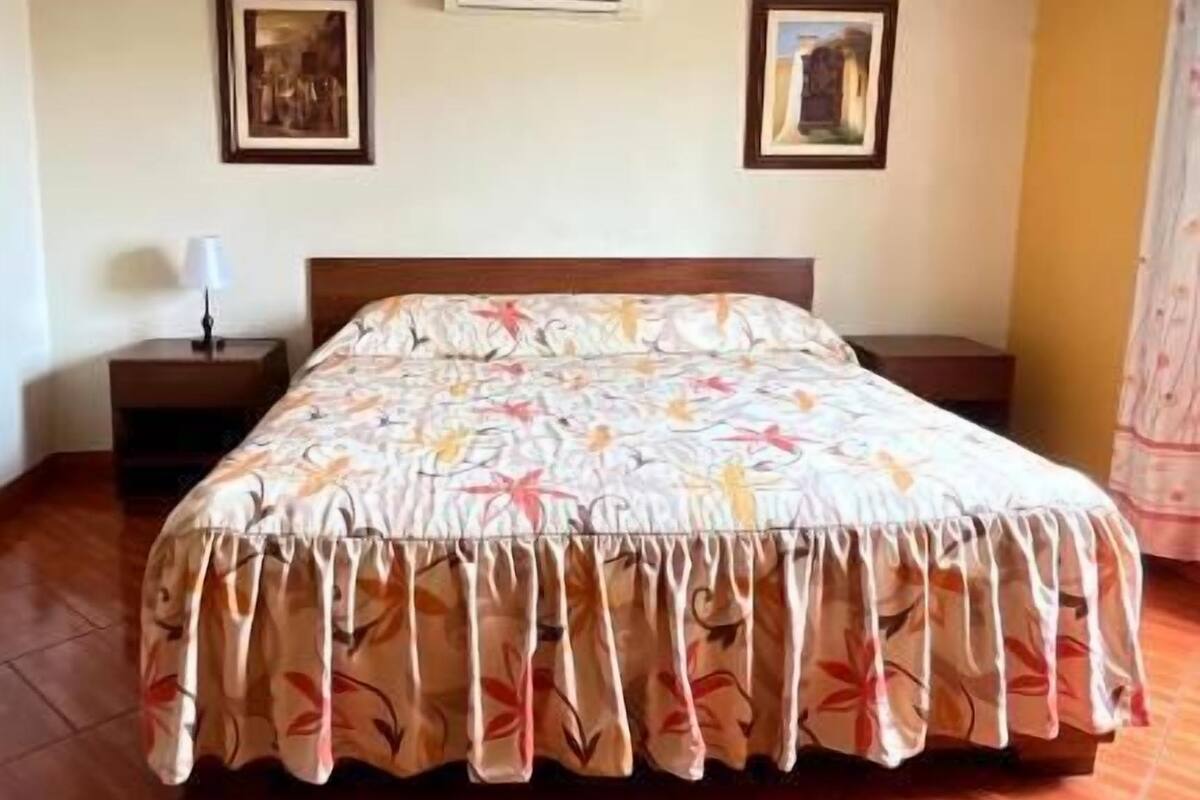 Un dormitorio con cama, mesita de noche, aire acondicionado y dos cuadros en la pared.
