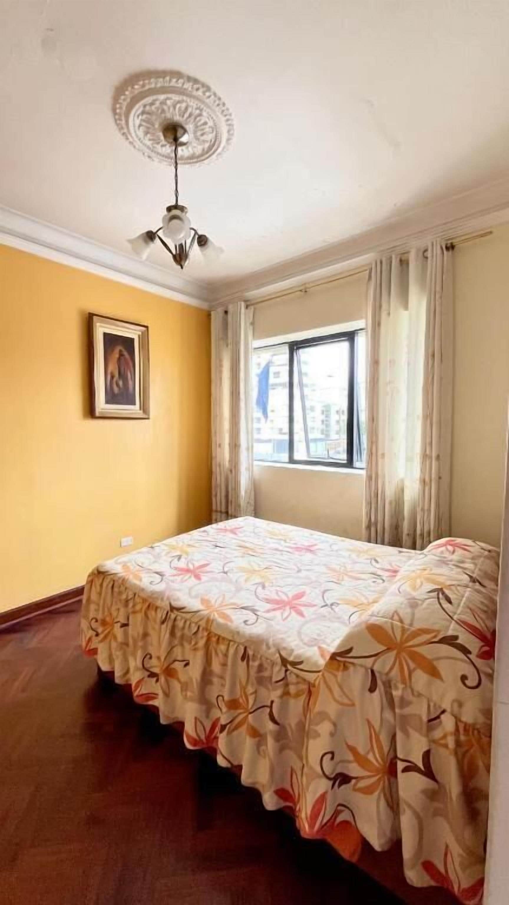 Una stanza con un letto, un ventilatore a soffitto, una finestra con tende e un quadro appeso al muro.