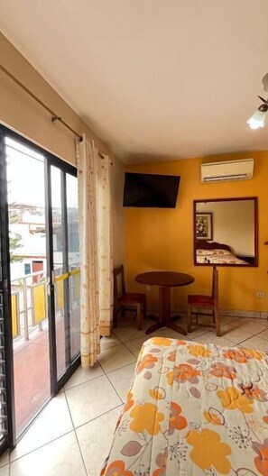 Habitación de hotel con una cama, una mesita con sillas, un televisor montado en la pared y un balcón con puerta corrediza.