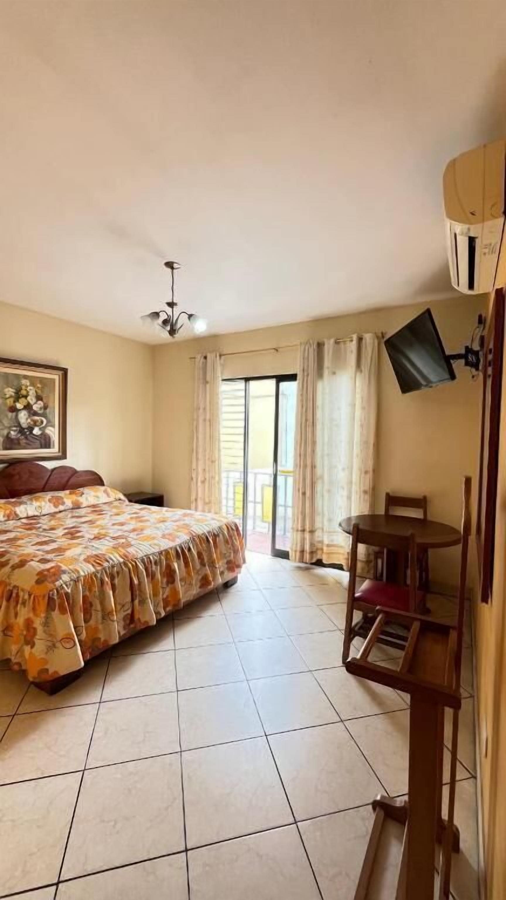 Habitación de hotel con cama, televisión montada en la pared, una mesita y balcón con cortinas.