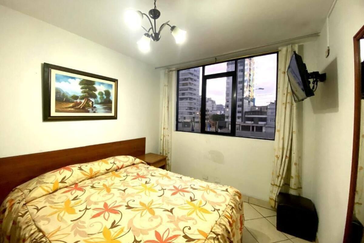 Un dormitorio con cama, un cuadro, una ventana con vista a la ciudad, una lámpara en el techo y un televisor montado en la pared.