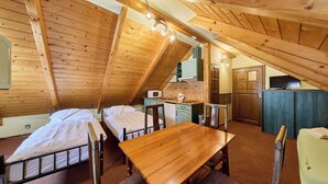 Um quarto de sótão pequeno com duas camas, uma mesa de madeira, uma cadeira, um espaço de cozinha e uma televisão.