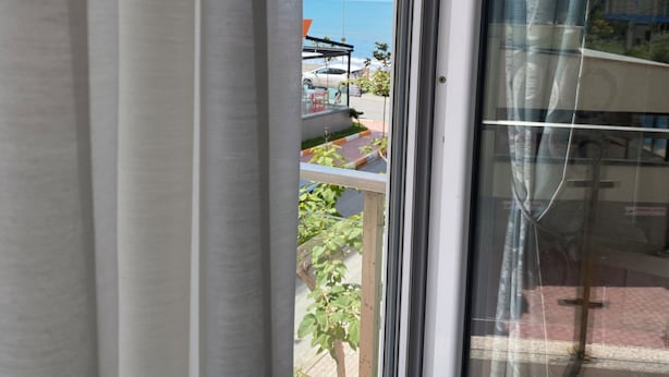 Vista a través de una ventana con cortina, cielo despejado y un atisbo de un edificio y vegetación.