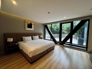Une chambre moderne avec un grand lit, une table de chevet, une lampe fixée au mur et une fenêtre donnant sur un paysage verdoyant.