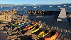 Una playa rocosa con kayaks, un muelle y embarcaciones al fondo.