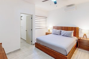 Ein Schlafzimmer mit hölzernem Bettgestell, Deckenventilator, Fenster mit Jalousien und Nachttisch mit Lampe.