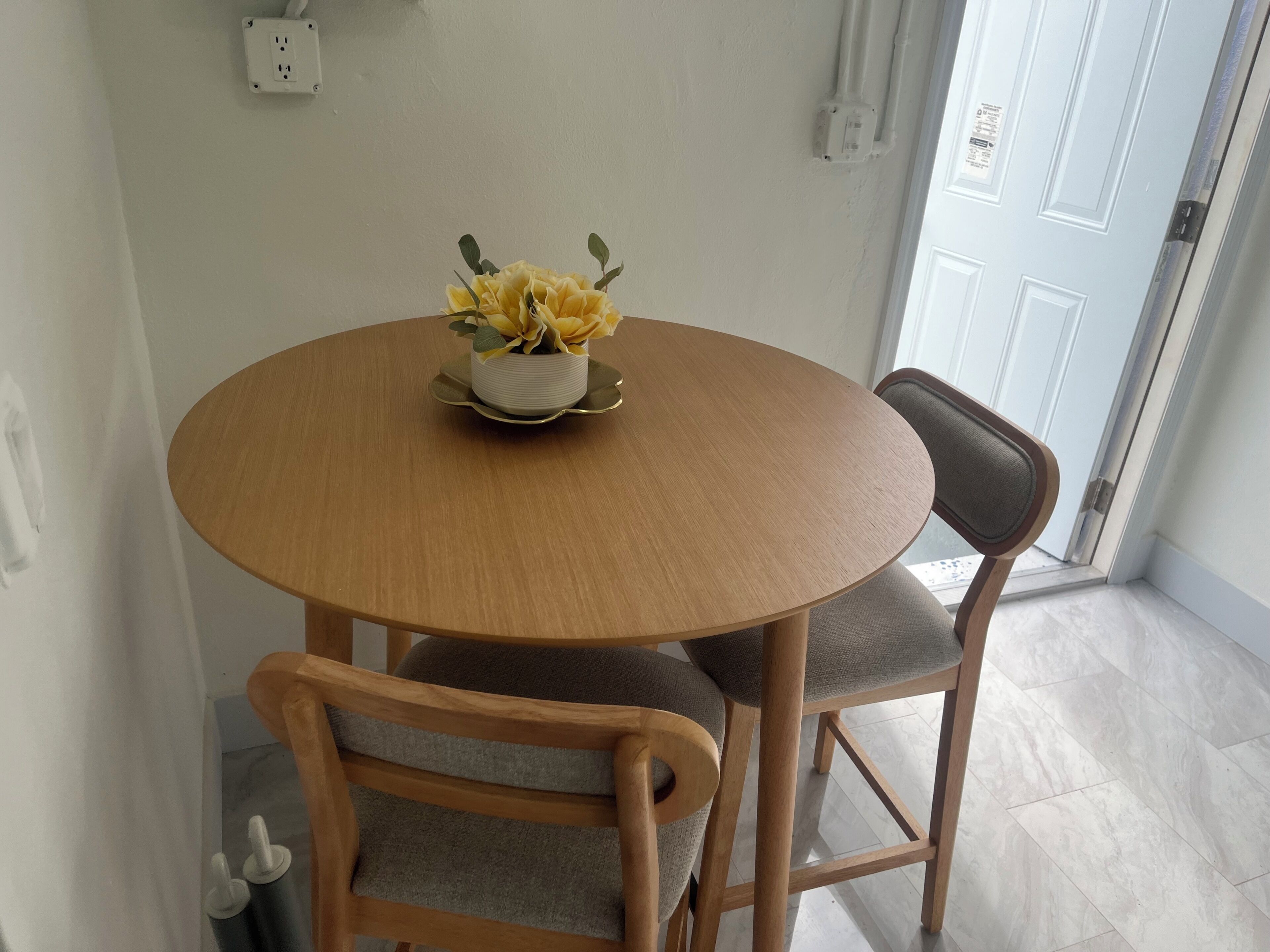 Una mesa de madera redonda con un jarrón blanco lleno de flores amarillas, dos sillas grises tapizadas y una puerta blanca al fondo.