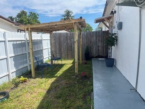 A backyard with a wooden pergola, two blue chairs, a potted plant, and a concrete pathway. - Fabulous studio with AC in vibrant Miami Gardens (Miami Gardens)