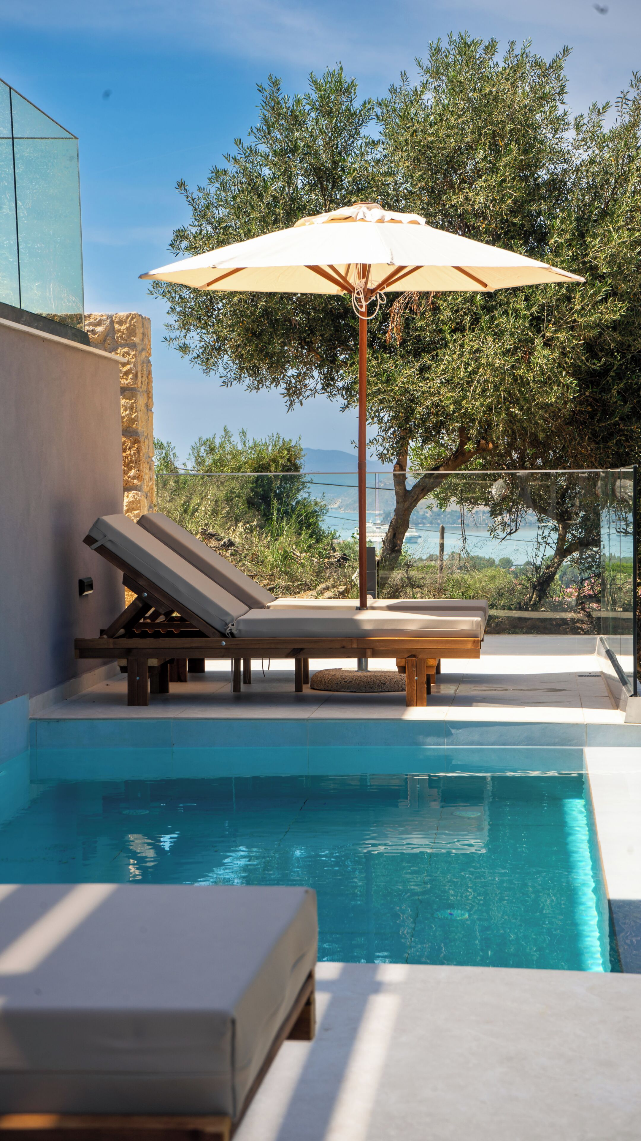 Zona de piscina con sillones de descanso y una sombrilla, con vista a una masa de agua y vegetación.