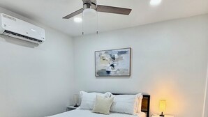 Un dormitorio con una cama, ventilador de techo, aire acondicionado, lámpara de noche y un cuadro en la pared.