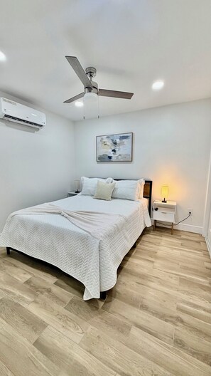 Un dormitorio con una cama, ventilador de techo, aire acondicionado, lámpara de noche y un cuadro en la pared.