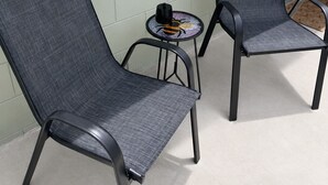 Dos sillas de malla gris con estructura metálica, una mesita redonda con un objeto negro encima y una pared blanca con un tono verdoso.