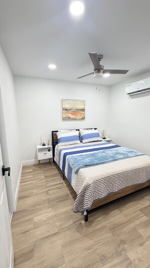A bedroom with a bed, nightstands, a ceiling fan, an air conditioning unit, and a painting on the wall.