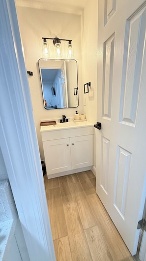 A bathroom with a white vanity, a mirror with lights, a wooden floor, and a white door.