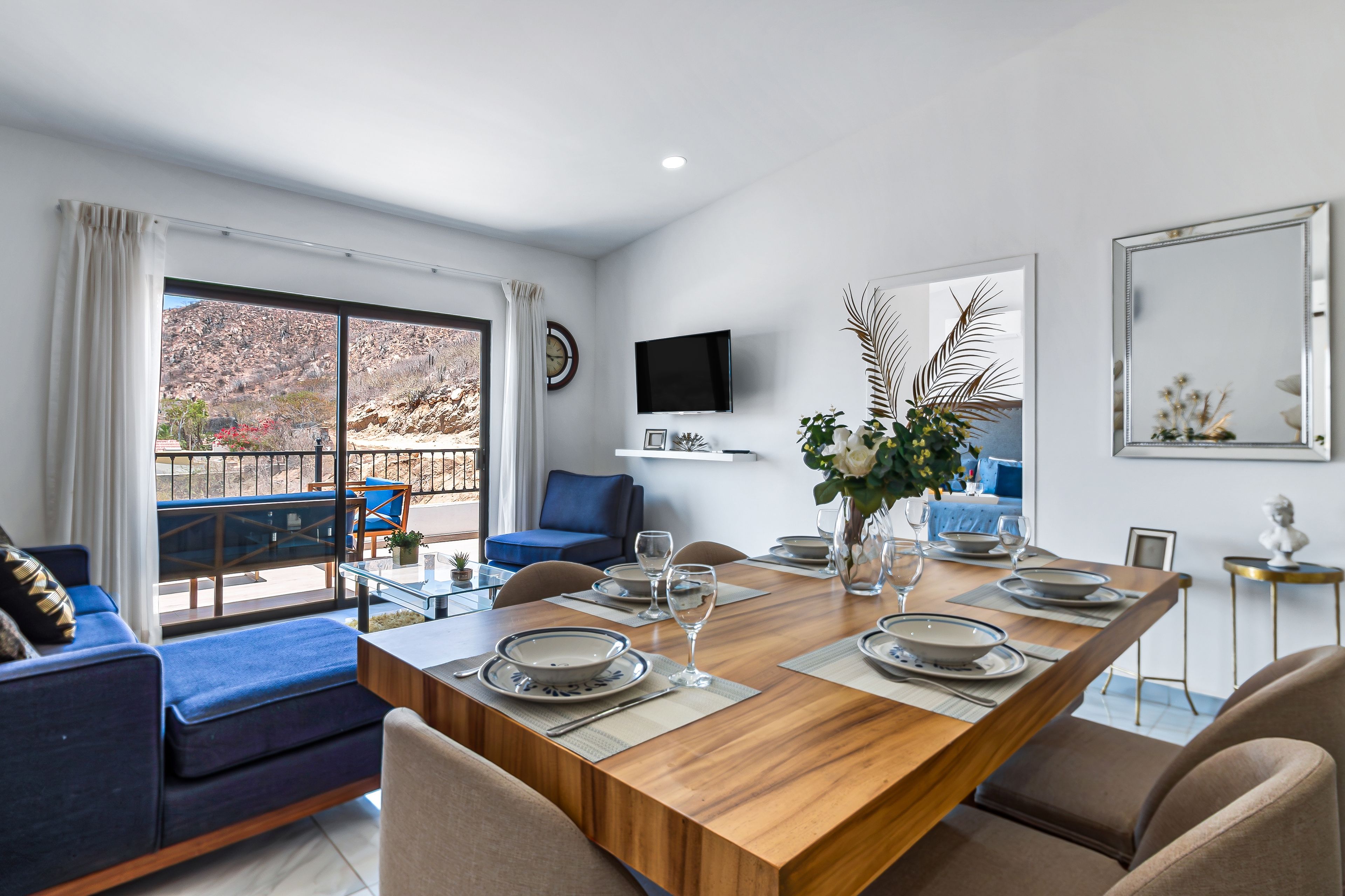 Een eetruimte met een houten tafel gedekt voor vier personen, omringd door blauwe banken en beige fauteuils. Een televisie is aan de muur bevestigd, en een groot raam biedt uitzicht op het omliggende landschap.
