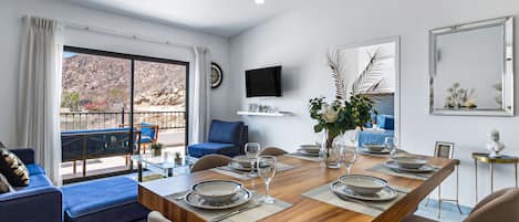 A dining area with a wooden table set for four, surrounded by blue sofas and beige armchairs. A TV is mounted on the wall, and a large window offers a view of the outdoors.