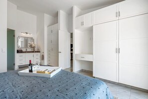 Quarto com cama, mesa de cabeceira com garrafa e copos, espelho e armários brancos.