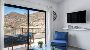 A modern living room with a glass coffee table, a blue sofa, a flat-screen TV, and a balcony with a view of a mountainous landscape. - Morgan Residences 607a @ Boutique Hotel With Pool, Gym, Spa (Cabo San Lucas)