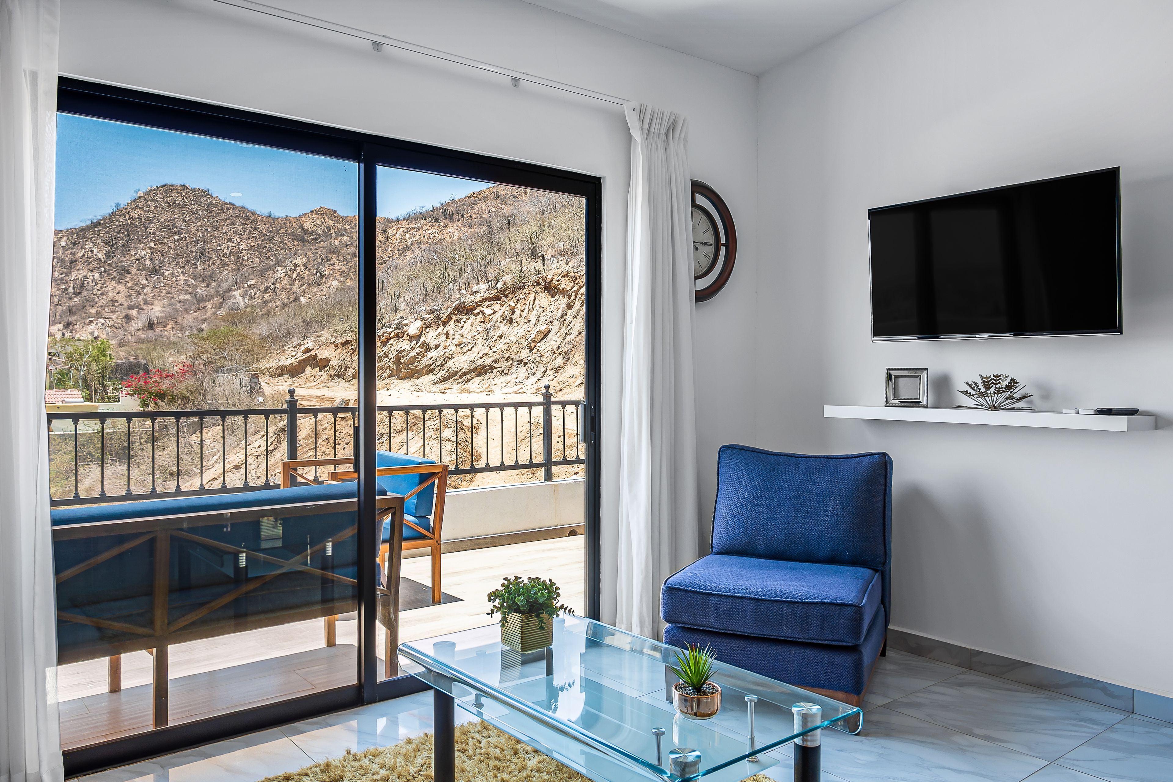 A modern living room with a glass coffee table, a blue sofa, and a TV mounted on the wall.